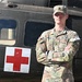 A U.S. Army Sergeant named Whitley stands with arms crossed in front of a military medical helicopter marked with a Red Cross symbol. He is wearing Operational Camouflage Pattern (OCP) uniform with visible rank insignia, name tape, U.S. Army tape, a unit patch, and a 'Competitor 4' armband. The image appears to be an official or promotional military photograph.