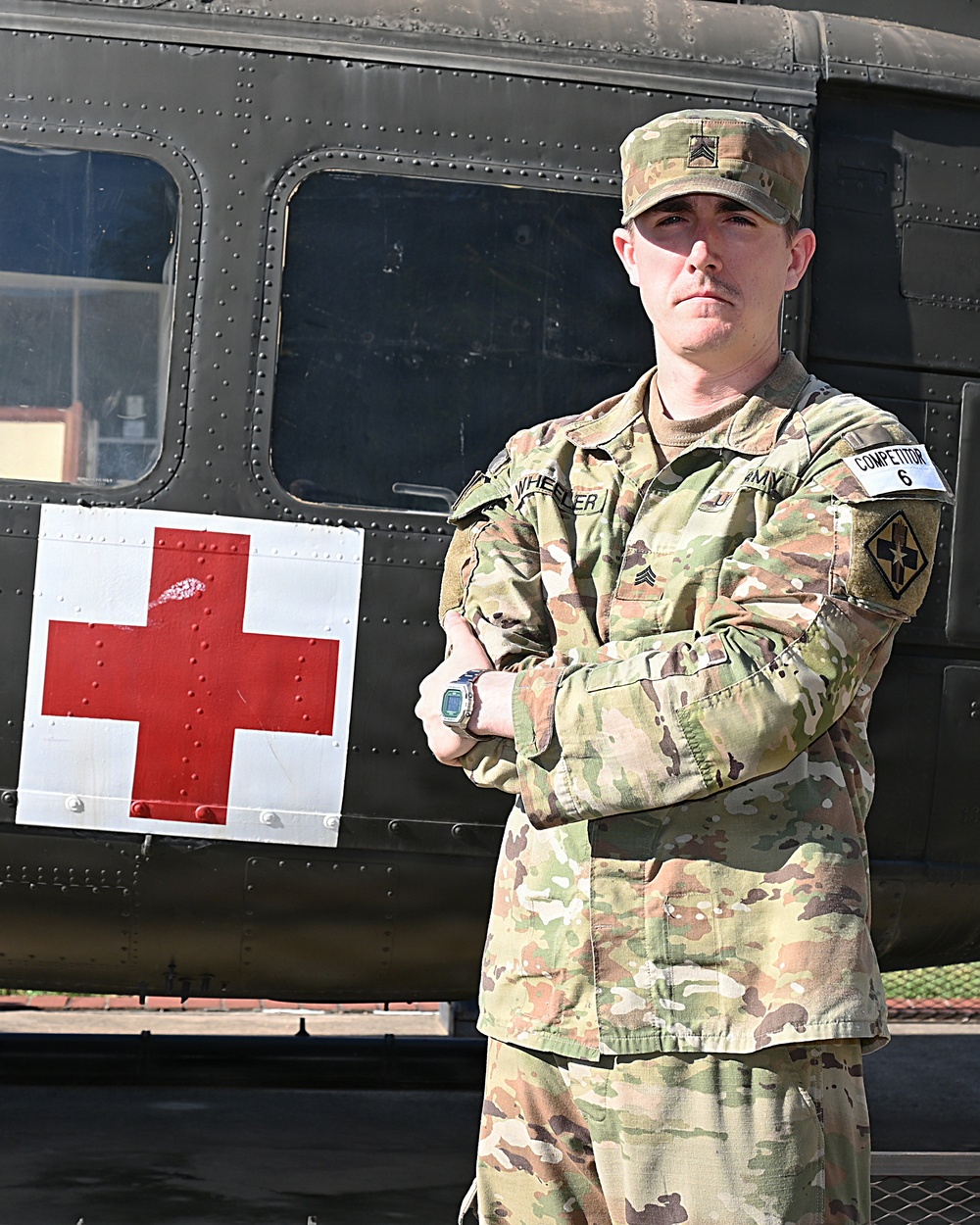A U.S. Army Sergeant in Operational Camouflage Pattern (OCP) uniform stands with arms crossed in front of a military medical vehicle marked with a Red Cross symbol. The soldier wears a name tape reading 'WHEELER' and an 'ARMY' tape, along with a 'COMPETITOR 6' armband on the right sleeve. The image appears to be an official or semi-official portrait-style photograph.