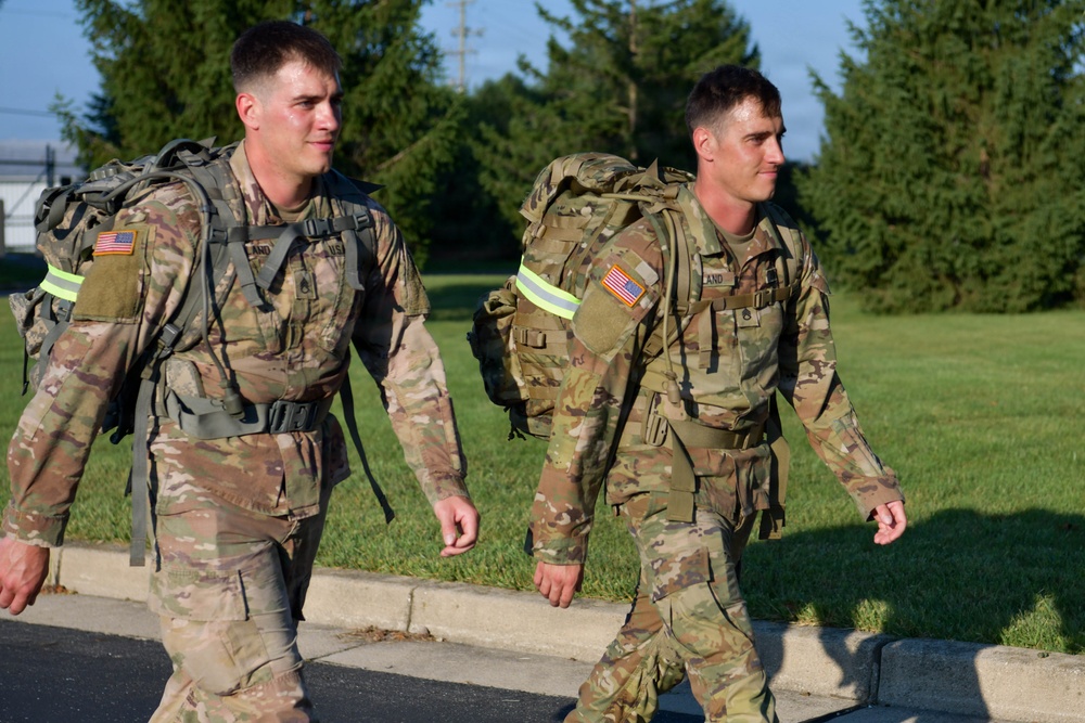 Two U.S. Army soldiers in MultiCam (OCP) camouflage uniforms are conducting a ruck march along a paved road. Both are carrying large military rucksacks equipped with high-visibility reflective bands, suggesting a safety-compliant road march. The setting appears to be a military installation with manicured grass and tree lines in the background.