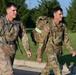 Two U.S. Army soldiers in MultiCam (OCP) camouflage uniforms are conducting a ruck march along a paved road. Both are carrying large military rucksacks equipped with high-visibility reflective bands, suggesting a safety-compliant road march. The setting appears to be a military installation with manicured grass and tree lines in the background.