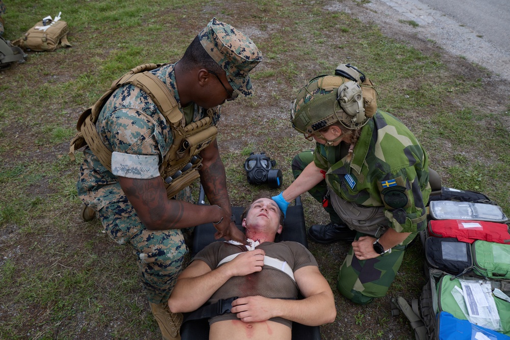Two military personnel from different nations are providing tactical combat casualty care (TCCC) to a simulated casualty lying on the ground. One individual wears US Marine Corps MARPAT digital camouflage with a plate carrier, while the other wears Swedish Armed Forces M90 camouflage with a tactical helmet bearing a Swedish flag patch. This appears to be a joint military medical training exercise.