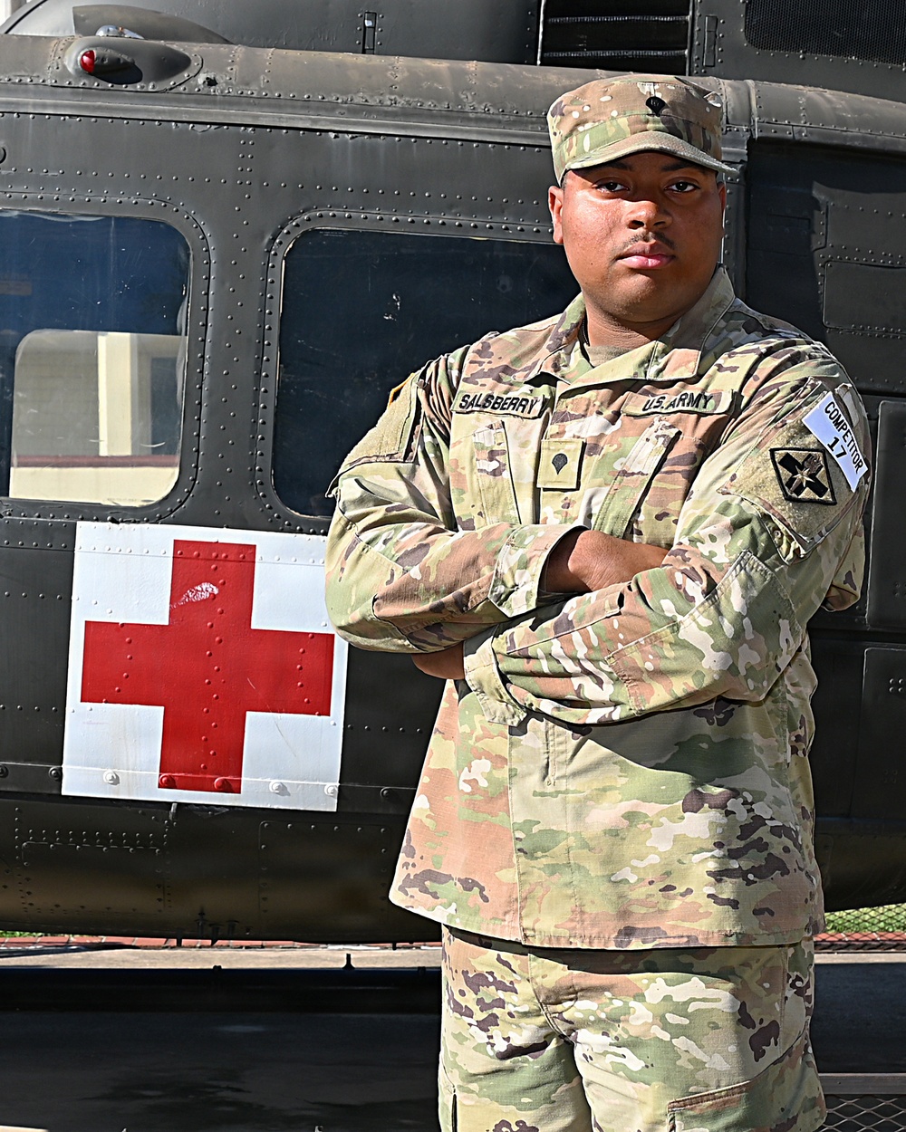 A U.S. Army soldier named Salsberry stands with arms crossed in front of a military medical helicopter marked with a red cross symbol. The soldier is wearing OCP (Operational Camouflage Pattern) uniform with visible name tape, U.S. Army tape, and a unit patch on the left sleeve. A 'Competitor 17' tab is visible on the right sleeve, suggesting this may be a competition or best warrior event photo.
