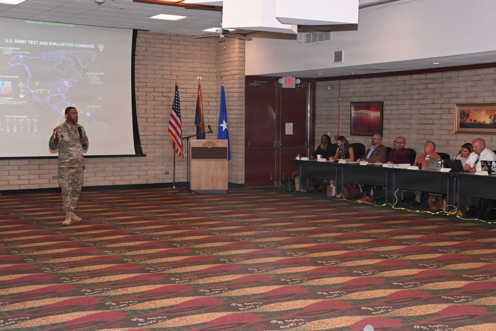A U.S. Army soldier in combat uniform is delivering a briefing to a panel of civilian and military attendees seated at a long table. The presentation slide visible on the projection screen reads 'U.S. Army Test and Evaluation Command' and includes a map. The setting appears to be a conference or meeting room with patterned carpet, brick walls, and flags displayed.