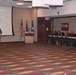 A U.S. Army soldier in combat uniform is delivering a briefing to a panel of civilian and military attendees seated at a long table. The presentation slide visible on the projection screen reads 'U.S. Army Test and Evaluation Command' and includes a map. The setting appears to be a conference or meeting room with patterned carpet, brick walls, and flags displayed.