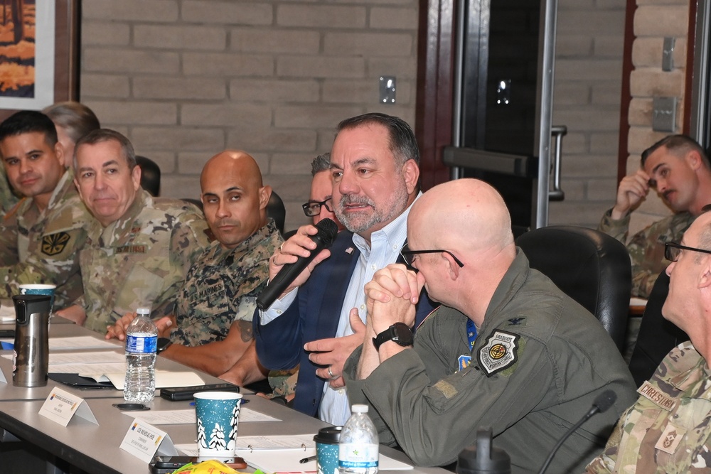 A formal military briefing or roundtable meeting is taking place in an indoor conference room setting. A civilian man in a blue blazer is speaking into a handheld microphone, surrounded by uniformed military personnel from multiple branches including Army, Marines, and Air Force. Name placards are visible on the table, suggesting an official organized meeting.