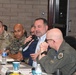 A formal military briefing or roundtable meeting is taking place in an indoor conference room setting. A civilian man in a blue blazer is speaking into a handheld microphone, surrounded by uniformed military personnel from multiple branches including Army, Marines, and Air Force. Name placards are visible on the table, suggesting an official organized meeting.