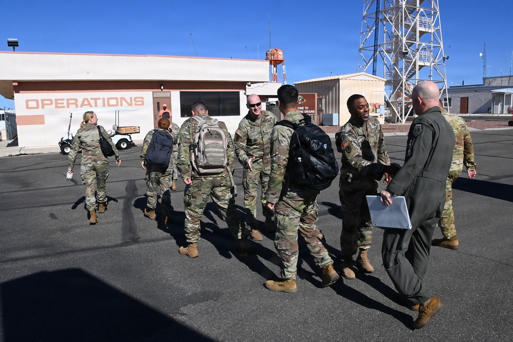A group of military personnel in OCP (Operational Camouflage Pattern) uniforms are gathered outside a building labeled 'OPERATIONS' on what appears to be a military installation or airfield. Several soldiers are carrying backpacks and appear to be arriving or departing, greeted by a person in a flight suit holding a clipboard or folder. The setting includes a communications or observation tower in the background under clear blue skies.
