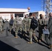 A group of military personnel in OCP (Operational Camouflage Pattern) uniforms are gathered outside a building labeled 'OPERATIONS' on what appears to be a military installation or airfield. Several soldiers are carrying backpacks and appear to be arriving or departing, greeted by a person in a flight suit holding a clipboard or folder. The setting includes a communications or observation tower in the background under clear blue skies.