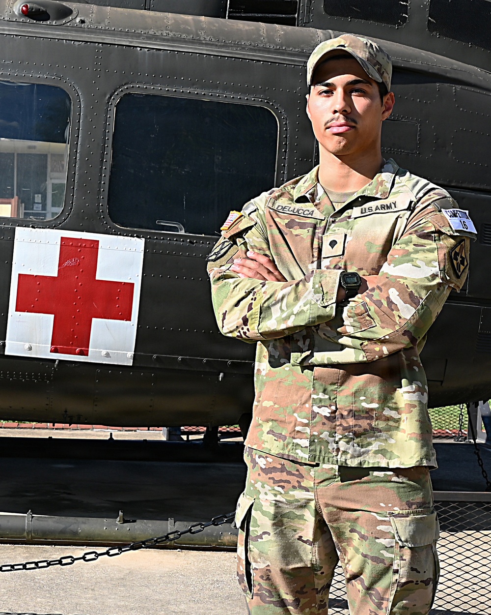 A U.S. Army soldier named DeLucca stands at attention with arms crossed in front of a military medical helicopter marked with a Red Cross symbol. The soldier is wearing OCP (Operational Camouflage Pattern) uniform with visible name tape, U.S. Army tape, and a competitor number tag (16) on the left sleeve. The image appears to be an official or promotional military photograph.