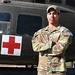 A U.S. Army soldier named DeLucca stands at attention with arms crossed in front of a military medical helicopter marked with a Red Cross symbol. The soldier is wearing OCP (Operational Camouflage Pattern) uniform with visible name tape, U.S. Army tape, and a competitor number tag (16) on the left sleeve. The image appears to be an official or promotional military photograph.