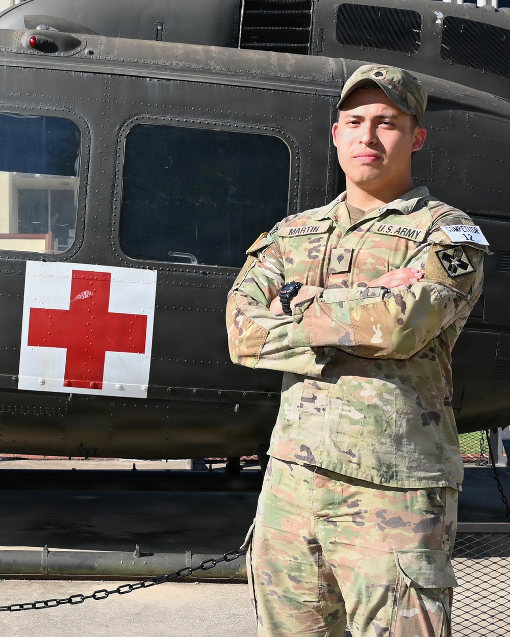 A U.S. Army soldier named Martin stands at attention with arms crossed in front of a dark olive-green military medical helicopter marked with a Red Cross symbol. The soldier is wearing OCP (Operational Camouflage Pattern) uniform with a 'COMPETITOR 12' tag visible on the right sleeve, suggesting participation in a military competition. The setting appears to be an outdoor military installation or event.