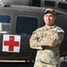 A U.S. Army soldier named Martin stands at attention with arms crossed in front of a dark olive-green military medical helicopter marked with a Red Cross symbol. The soldier is wearing OCP (Operational Camouflage Pattern) uniform with a 'COMPETITOR 12' tag visible on the right sleeve, suggesting participation in a military competition. The setting appears to be an outdoor military installation or event.