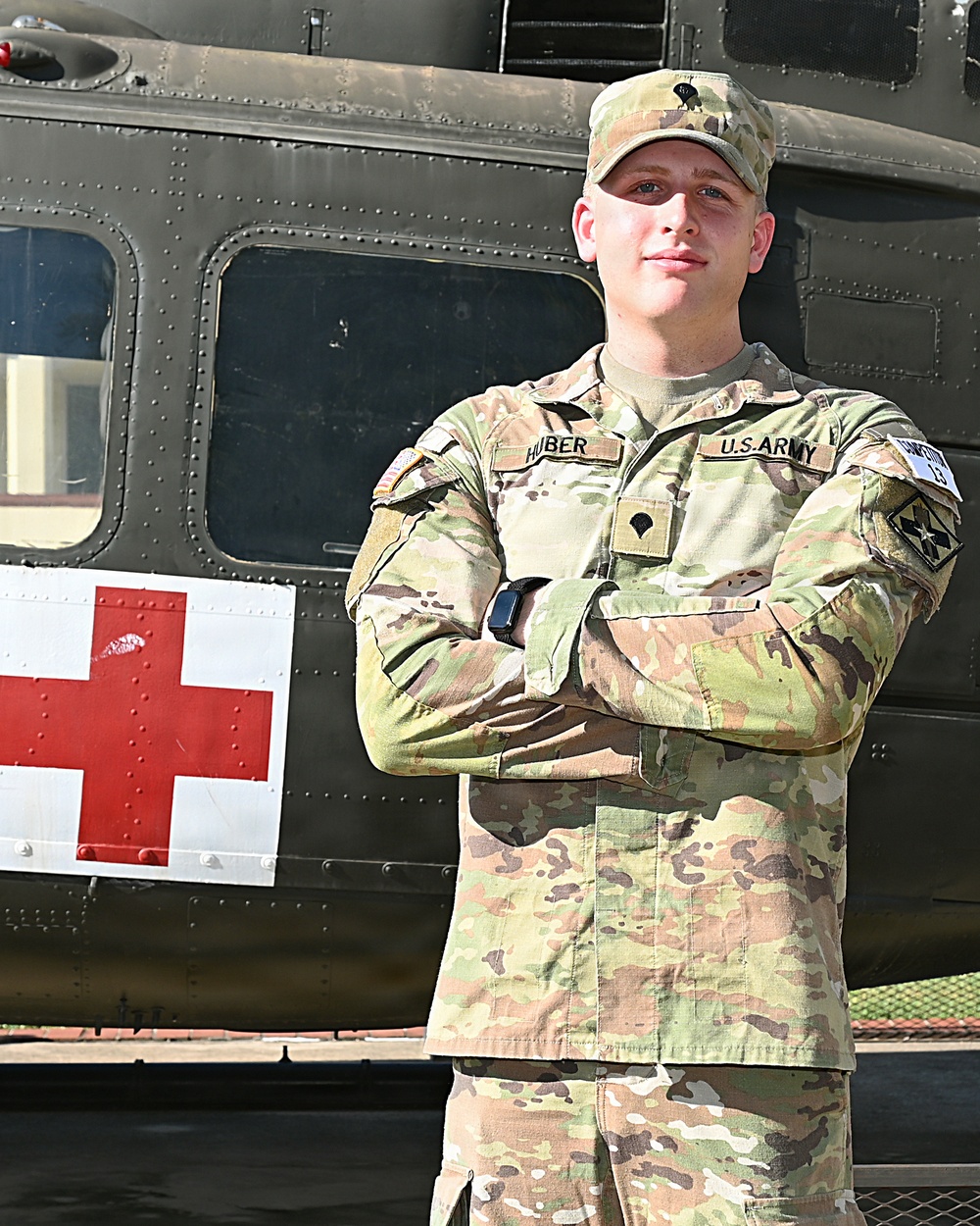 A U.S. Army soldier named Huber stands with arms crossed in front of a military medical helicopter marked with a red cross. He is wearing Operational Camouflage Pattern (OCP) uniform with visible rank insignia, name tape, and unit patches. The image appears to be an official or promotional military portrait taken outdoors in good lighting.