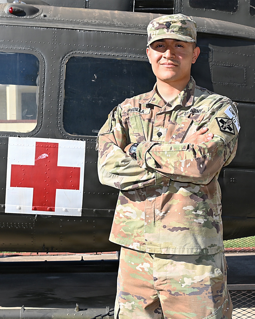 A U.S. Army soldier in Operational Camouflage Pattern (OCP) uniform stands with arms crossed in front of a military medical helicopter marked with a Red Cross symbol. The soldier's name tape reads 'PADILLA' and the right chest tape reads 'US ARMY'. A 'COMPETITOR 10' tab is visible on the left shoulder, and a unit patch is on the left sleeve.