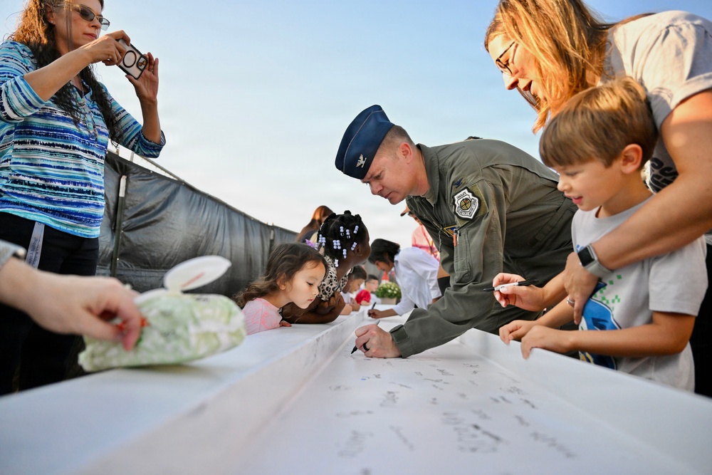 A military officer in a green flight suit and blue service cap is signing a large white banner or fabric alongside children and adults at what appears to be an outdoor community event. The scene captures a moment of interaction between a uniformed service member and civilian families, including young children. A woman in the background is photographing the event with a smartphone.