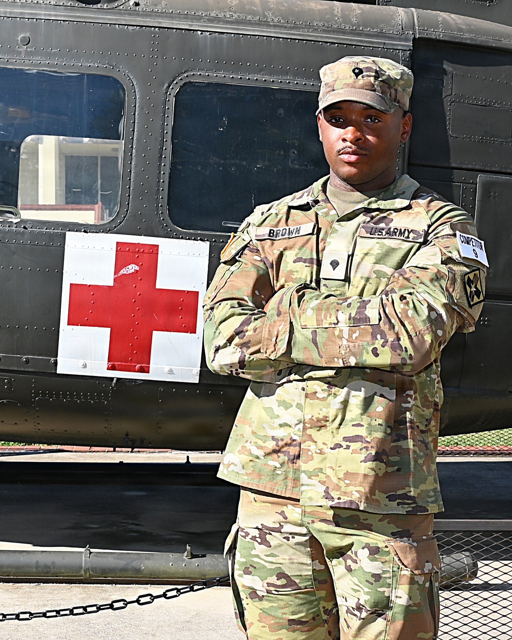 A U.S. Army soldier named Brown stands at attention with arms crossed in front of a military medical helicopter marked with a Red Cross symbol. The soldier is wearing Operational Camouflage Pattern (OCP) uniform with 'US ARMY' and 'BROWN' name tapes visible, along with unit patches. The setting appears to be an outdoor military installation or display area.