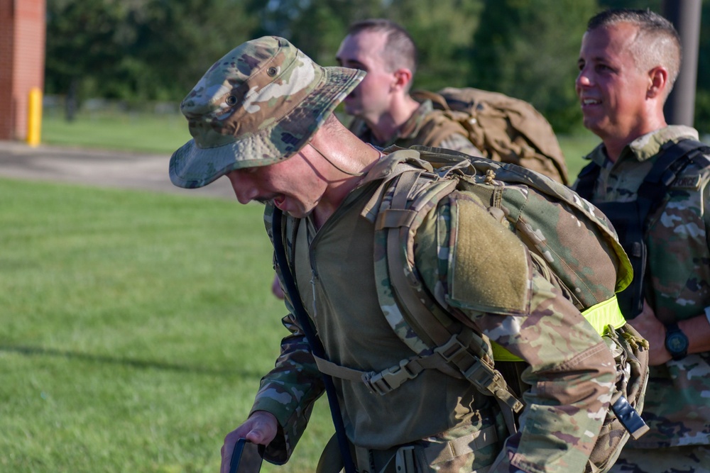 Three military personnel in Operational Camouflage Pattern (OCP) uniforms are engaged in what appears to be a ruck march or physical training exercise. The foreground soldier is bent forward under the weight of a heavy rucksack and appears to be exerting significant effort, while a second soldier in the background is smiling. A high-visibility yellow band is visible on the foreground soldier's rucksack.