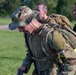 Three military personnel in Operational Camouflage Pattern (OCP) uniforms are engaged in what appears to be a ruck march or physical training exercise. The foreground soldier is bent forward under the weight of a heavy rucksack and appears to be exerting significant effort, while a second soldier in the background is smiling. A high-visibility yellow band is visible on the foreground soldier's rucksack.