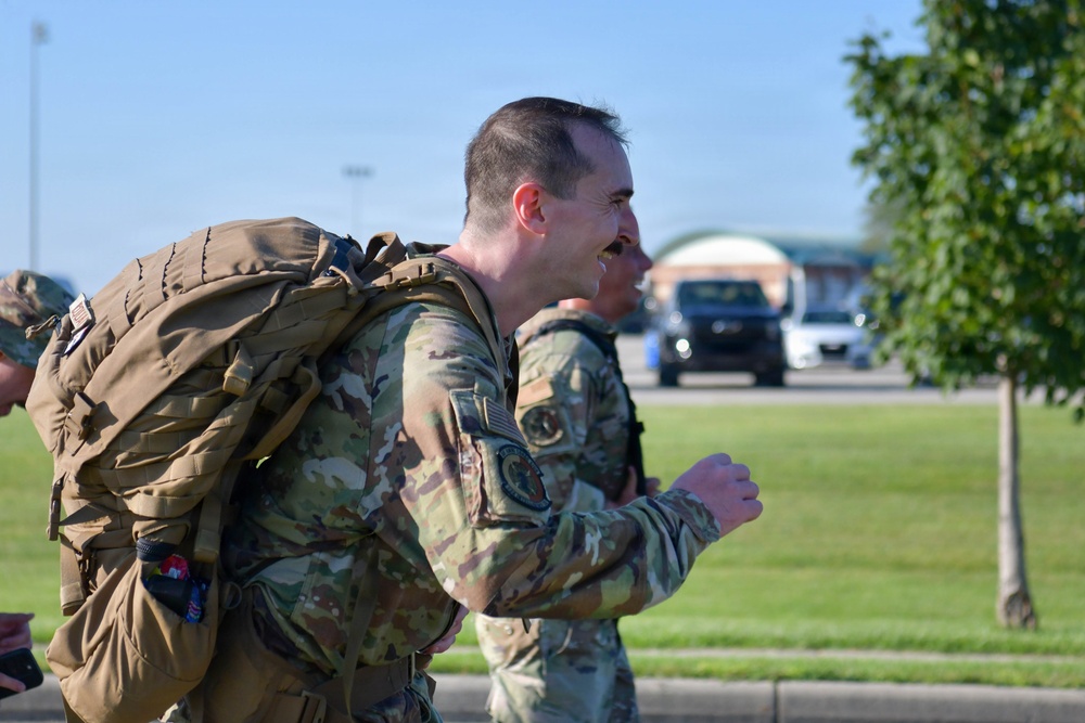 A military service member in OCP (Operational Camouflage Pattern) uniform is running or marching with a large tactical rucksack, smiling broadly. At least one additional service member in the same uniform is visible behind them. The setting appears to be a military installation based on the manicured grounds, parking area, and structures visible in the background.