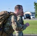 A military service member in OCP (Operational Camouflage Pattern) uniform is running or marching with a large tactical rucksack, smiling broadly. At least one additional service member in the same uniform is visible behind them. The setting appears to be a military installation based on the manicured grounds, parking area, and structures visible in the background.