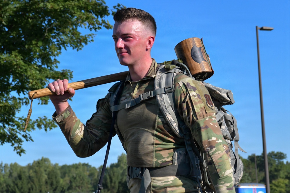 A young male soldier in US Army OCP (Operational Camouflage Pattern) uniform carries a large wooden mallet over his shoulder while wearing a military rucksack. His face shows signs of physical exertion, appearing flushed and sweaty, suggesting he is participating in a physically demanding military training event or competition. The outdoor setting features a clear blue sky and green trees in the background.
