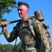 A young male soldier in US Army OCP (Operational Camouflage Pattern) uniform carries a large wooden mallet over his shoulder while wearing a military rucksack. His face shows signs of physical exertion, appearing flushed and sweaty, suggesting he is participating in a physically demanding military training event or competition. The outdoor setting features a clear blue sky and green trees in the background.