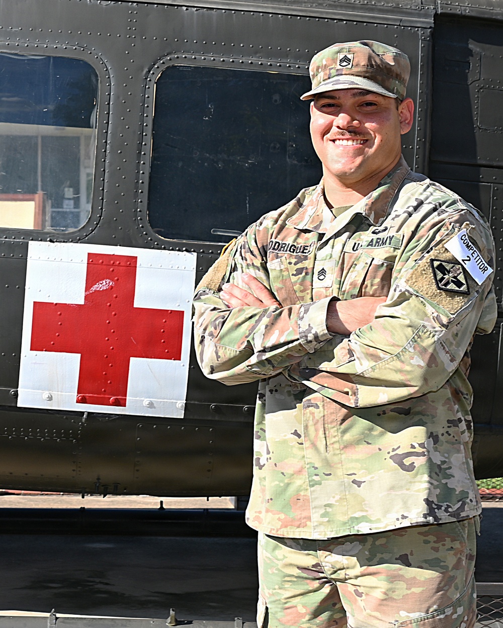 A U.S. Army Sergeant named Rodriguez stands at ease with arms crossed in front of a military medical vehicle marked with a Red Cross symbol. He is wearing Operational Camouflage Pattern (OCP) uniform with visible rank insignia, name tape, U.S. Army tape, a unit patch, and a 'Competitor 2' label. The image appears to be an official or promotional military photograph.