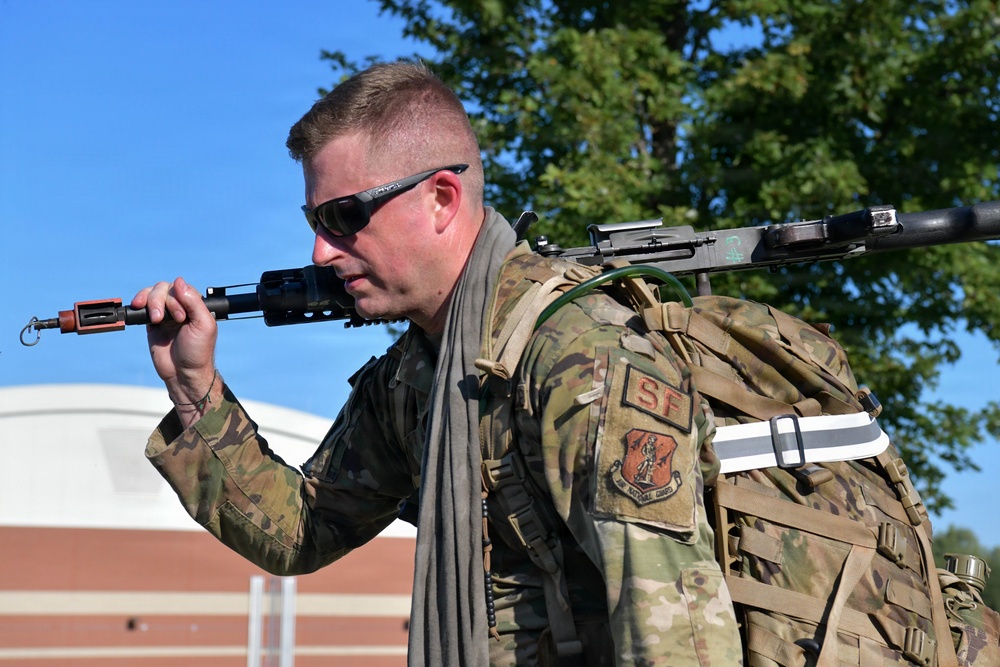 A male military service member wearing OCP (Operational Camouflage Pattern) uniform is carrying a large tactical backpack and holding a rifle over his shoulder. A patch on his left sleeve reads 'SF' indicating Air Force Security Forces. He is outdoors in what appears to be a base or institutional setting with trees and a brick building visible in the background.