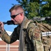 A male military service member wearing OCP (Operational Camouflage Pattern) uniform is carrying a large tactical backpack and holding a rifle over his shoulder. A patch on his left sleeve reads 'SF' indicating Air Force Security Forces. He is outdoors in what appears to be a base or institutional setting with trees and a brick building visible in the background.