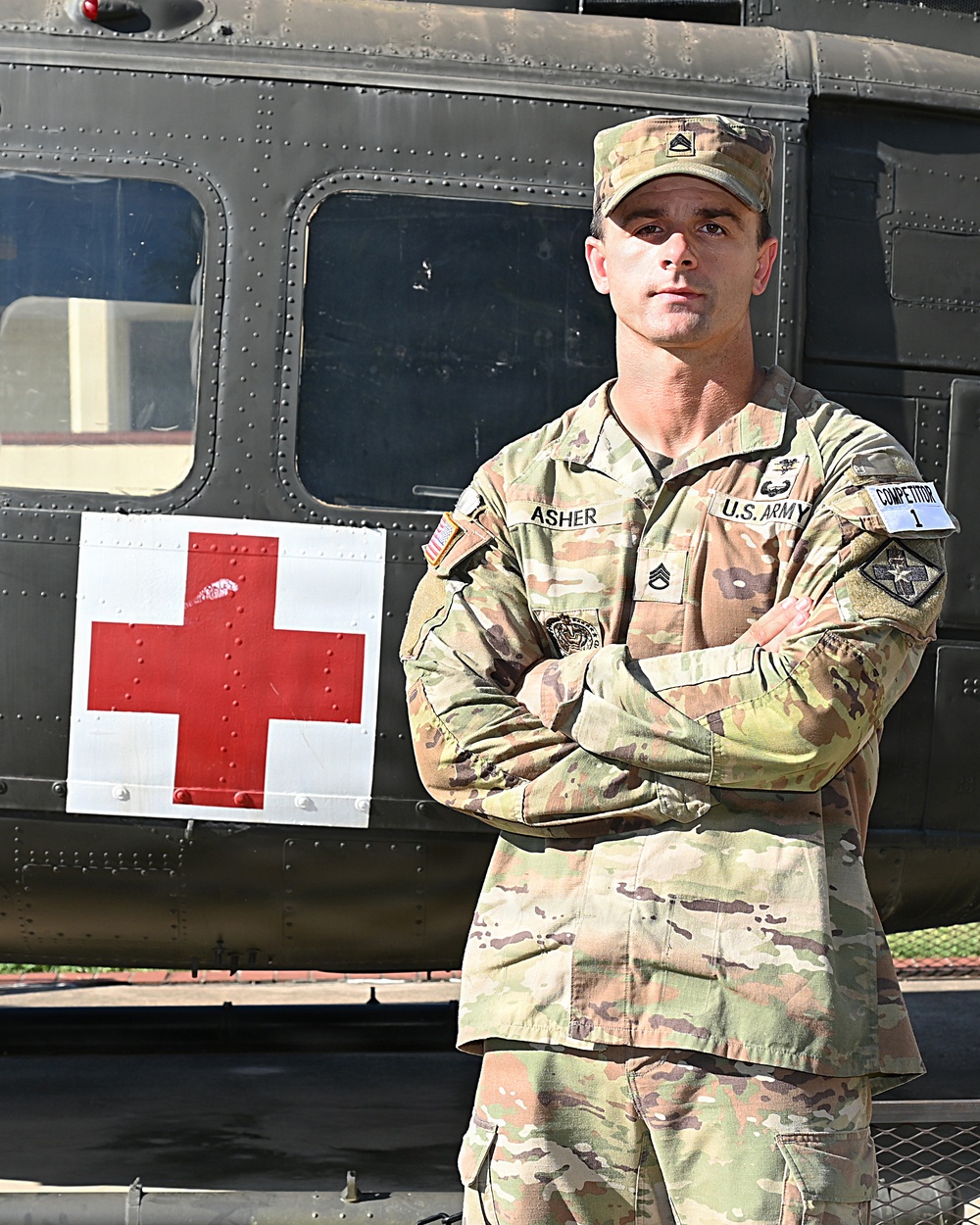 A U.S. Army Staff Sergeant named Asher stands at attention with arms crossed in front of a military medical helicopter marked with a Red Cross symbol. He is wearing OCP (Operational Camouflage Pattern) uniform with visible rank insignia, name tape, U.S. Army tape, unit patch, and a competition number tag labeled '1'. The image appears to be an official or promotional military photograph.