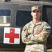 A U.S. Army Staff Sergeant named Asher stands at attention with arms crossed in front of a military medical helicopter marked with a Red Cross symbol. He is wearing OCP (Operational Camouflage Pattern) uniform with visible rank insignia, name tape, U.S. Army tape, unit patch, and a competition number tag labeled '1'. The image appears to be an official or promotional military photograph.