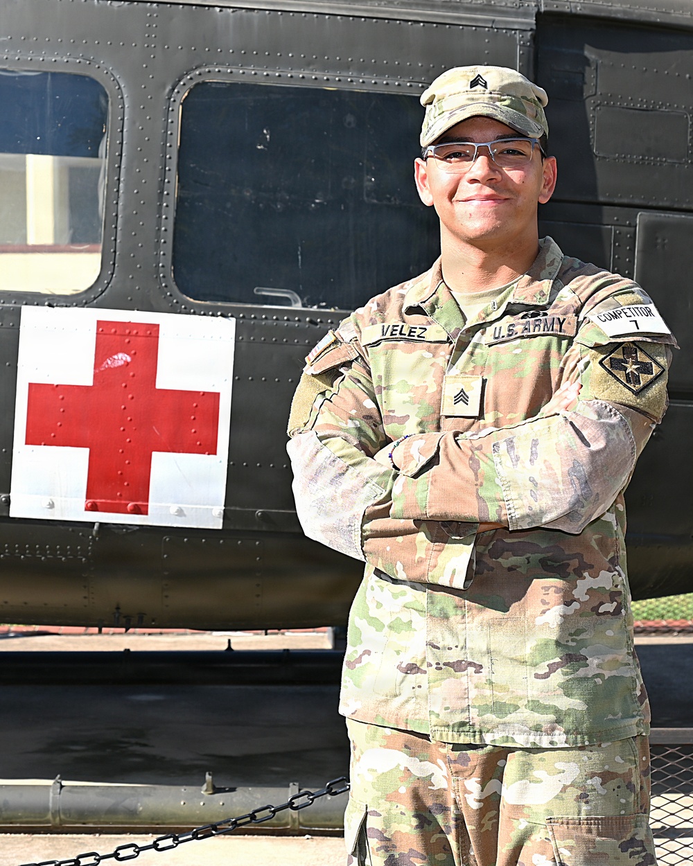A U.S. Army Sergeant named Velez stands with arms crossed in front of a military medical helicopter marked with a Red Cross symbol. The soldier is wearing Operational Camouflage Pattern (OCP) uniform with visible rank insignia, name tape, U.S. Army tape, and a unit patch. The image appears to be an official or promotional military photograph taken outdoors in daylight.
