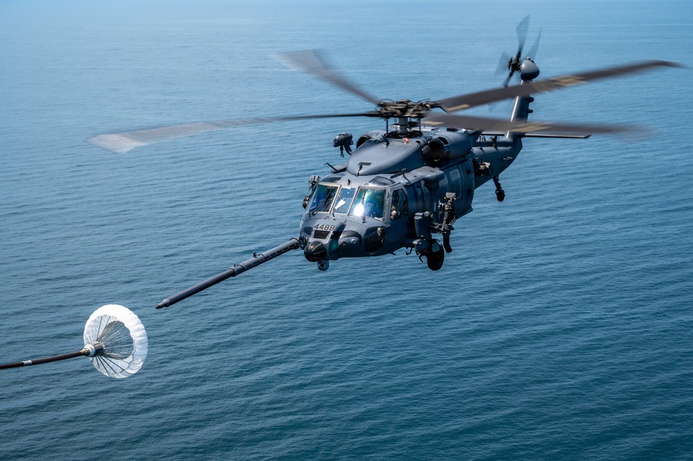 A military helicopter, marked with the number '4488', is captured in flight over open ocean water. The helicopter is equipped with an aerial refueling probe extended from its nose, approaching or connected to a drogue (basket) on a refueling hose. The image depicts an in-flight aerial refueling operation, likely conducted by a U.S. Navy or Air Force special operations helicopter.