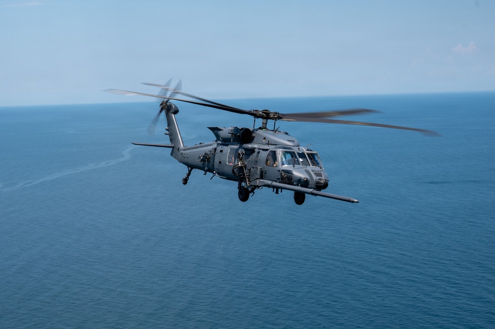 A military helicopter, consistent with an HH-60 Pave Hawk or MH-60 Black Hawk variant, is photographed in flight over a large body of open water. The aircraft is painted in a gray tactical finish and appears to be equipped with external fuel tanks and mission-specific equipment. The image is taken from another airborne platform at approximately the same altitude.