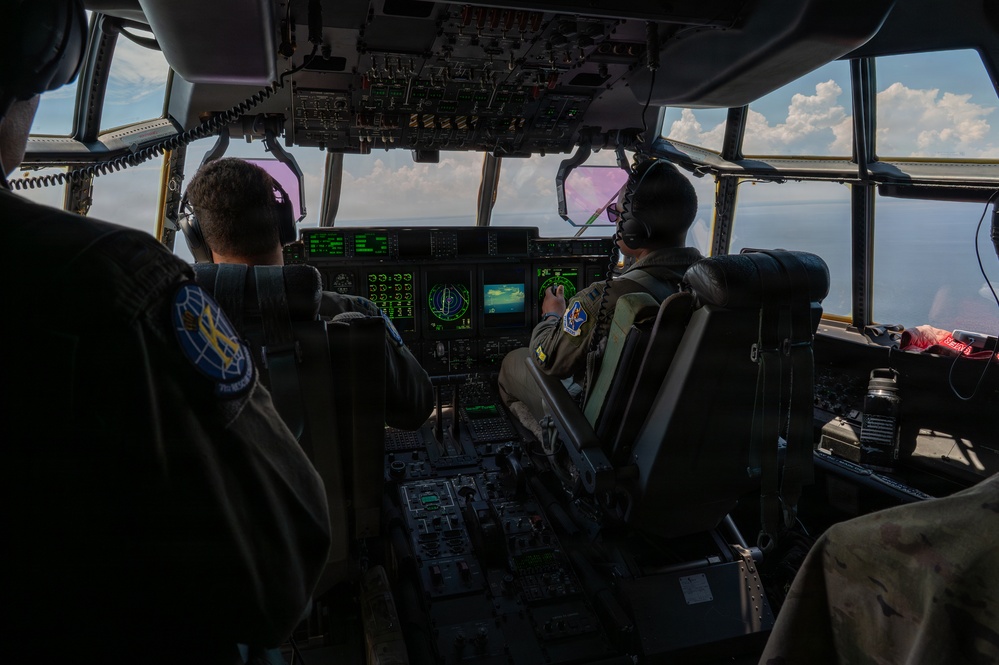 The image shows the interior cockpit of a large military transport aircraft, likely a C-130 or similar aircraft, with at least two crew members visible at their stations. The pilots are operating the aircraft in flight, with clouds and sky visible through the large multi-pane cockpit windows. Multiple avionics displays, instrument panels, and flight controls are visible throughout the cockpit.