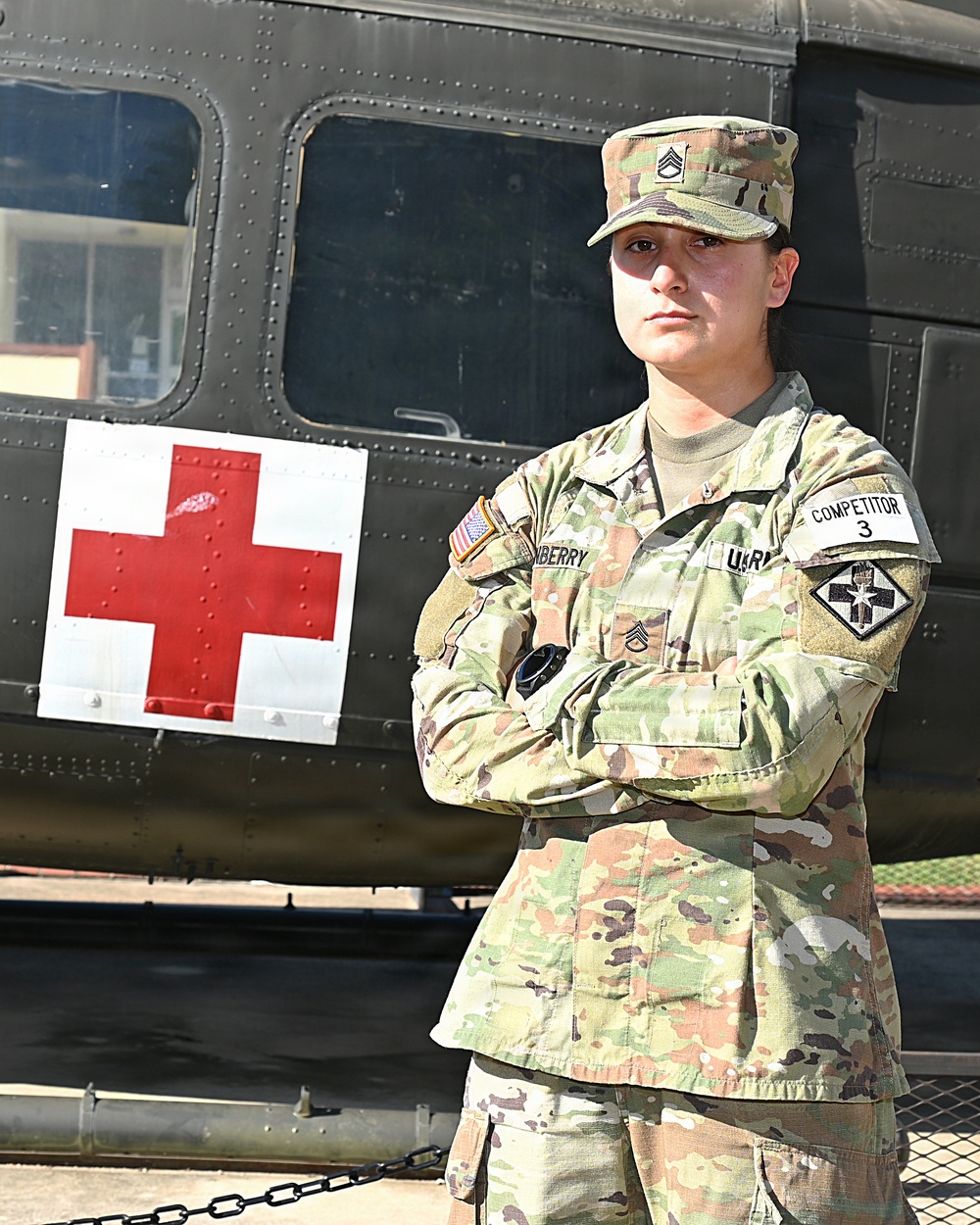 A U.S. Army soldier in Operational Camouflage Pattern (OCP) uniform stands at ease with arms crossed in front of a military medical helicopter marked with a Red Cross symbol. The soldier displays the rank of Staff Sergeant and wears a 'COMPETITOR 3' armband, suggesting participation in a military competition. The background features what appears to be a UH-60 Black Hawk or similar military medevac helicopter.