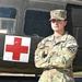 A U.S. Army soldier in Operational Camouflage Pattern (OCP) uniform stands at ease with arms crossed in front of a military medical helicopter marked with a Red Cross symbol. The soldier displays the rank of Staff Sergeant and wears a 'COMPETITOR 3' armband, suggesting participation in a military competition. The background features what appears to be a UH-60 Black Hawk or similar military medevac helicopter.