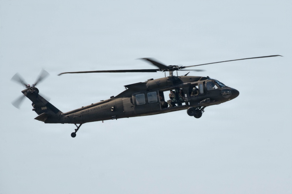 A United States Army UH-60 Black Hawk helicopter is captured in flight against an overcast sky. The aircraft is painted in olive drab/dark green military livery and has visible crew members aboard. The tail number '998' and 'UNITED STATES ARMY' text are legible on the fuselage.