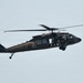 A United States Army UH-60 Black Hawk helicopter is captured in flight against an overcast sky. The aircraft is painted in olive drab/dark green military livery and has visible crew members aboard. The tail number '998' and 'UNITED STATES ARMY' text are legible on the fuselage.