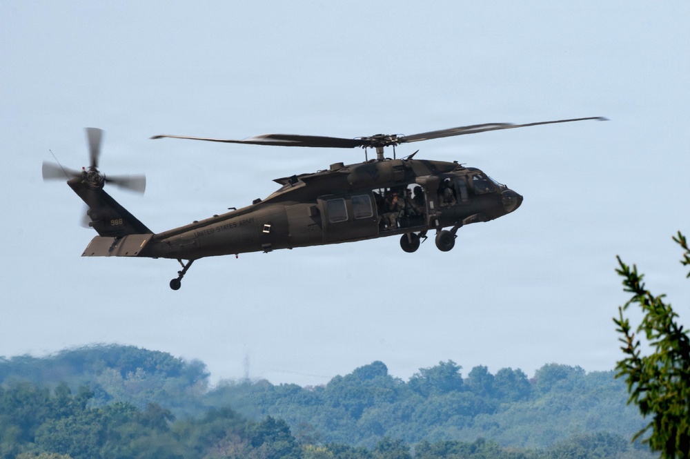 A United States Army UH-60 Black Hawk helicopter is captured in low-altitude flight against a pale blue sky. The aircraft bears the tail number '988' and 'UNITED STATES ARMY' markings on the fuselage. Personnel are visible through the open cabin door, and the helicopter appears to be in a hover or slow-flight maneuver over a forested landscape.