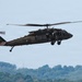 A United States Army UH-60 Black Hawk helicopter is captured in low-altitude flight against a pale blue sky. The aircraft bears the tail number '988' and 'UNITED STATES ARMY' markings on the fuselage. Personnel are visible through the open cabin door, and the helicopter appears to be in a hover or slow-flight maneuver over a forested landscape.