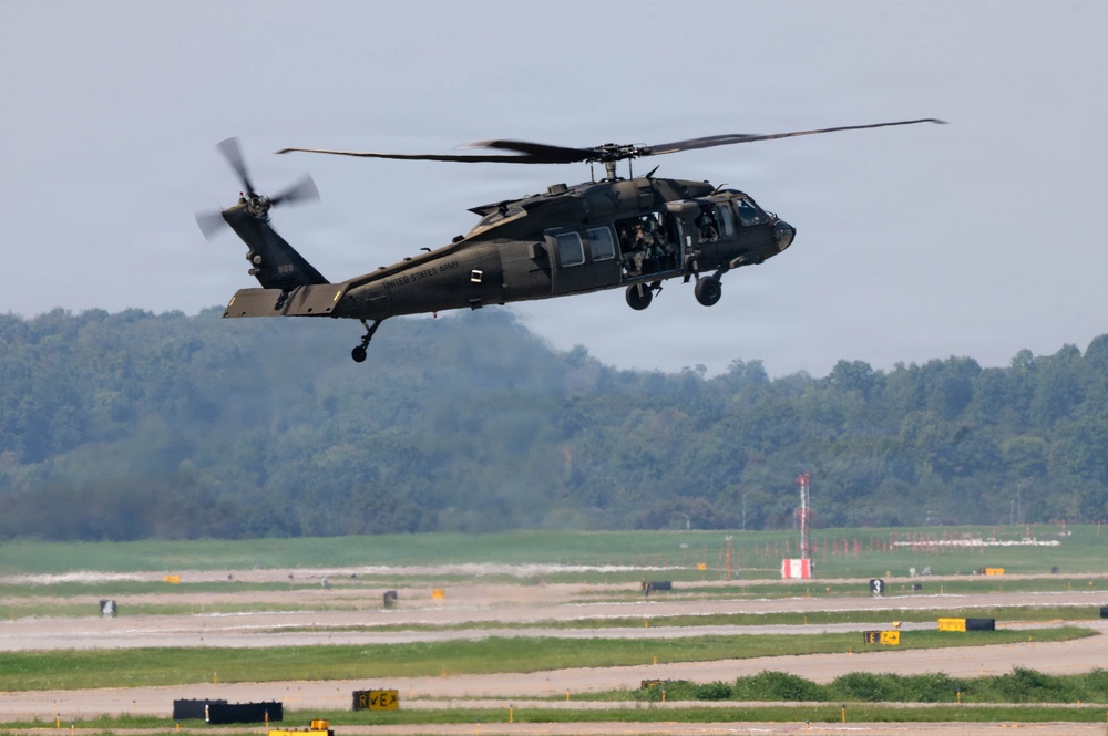 A United States Army UH-60 Black Hawk helicopter is captured in low-altitude flight over what appears to be a military airfield or airstrip. The helicopter is painted in standard olive drab/dark green military livery with 'UNITED STATES ARMY' markings visible on the fuselage. The main and tail rotors are in motion, and at least one crew member is visible through the open side door.