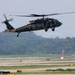 A United States Army UH-60 Black Hawk helicopter is captured in low-altitude flight over what appears to be a military airfield or airstrip. The helicopter is painted in standard olive drab/dark green military livery with 'UNITED STATES ARMY' markings visible on the fuselage. The main and tail rotors are in motion, and at least one crew member is visible through the open side door.