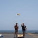 Two young men are playing cornhole on what appears to be the flight deck of a naval vessel, with open ocean visible in the background. One player has just released a beige bean bag, which is captured mid-air, while the other holds additional bags and watches. The setting is a large flat deck surface with painted markings typical of a military ship.