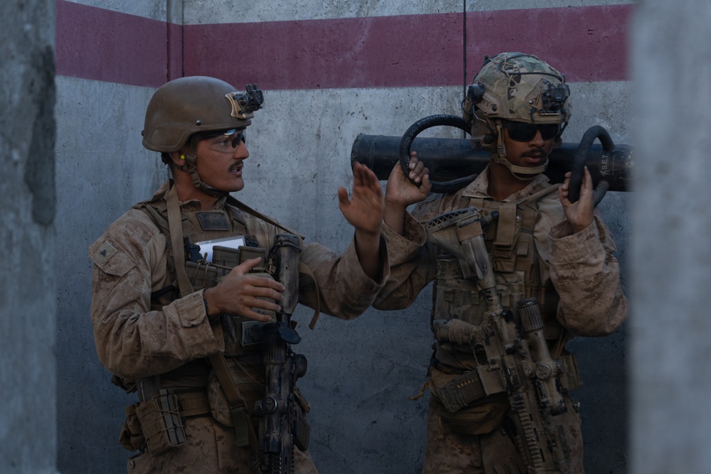 Two military personnel in desert camouflage uniforms are positioned against a concrete wall with red and gray painted stripes. They appear to be in active communication or coordination, with one soldier gesturing with open hands. Both are fully equipped with tactical gear, helmets, weapons, and body armor.