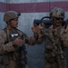Two military personnel in desert camouflage uniforms are positioned against a concrete wall with red and gray painted stripes. They appear to be in active communication or coordination, with one soldier gesturing with open hands. Both are fully equipped with tactical gear, helmets, weapons, and body armor.