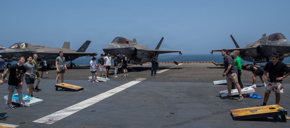 Military personnel are playing cornhole (bean bag toss) on the flight deck of a naval vessel, likely an amphibious assault ship. Multiple F-35B Lightning II stealth fighter jets are parked in the background on the deck. The scene appears to be a morale or recreational event held during a deployment, with the open ocean visible in the background.
