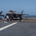 Military personnel are playing cornhole (bean bag toss) on the flight deck of a naval vessel, likely an amphibious assault ship. Multiple F-35B Lightning II stealth fighter jets are parked in the background on the deck. The scene appears to be a morale or recreational event held during a deployment, with the open ocean visible in the background.