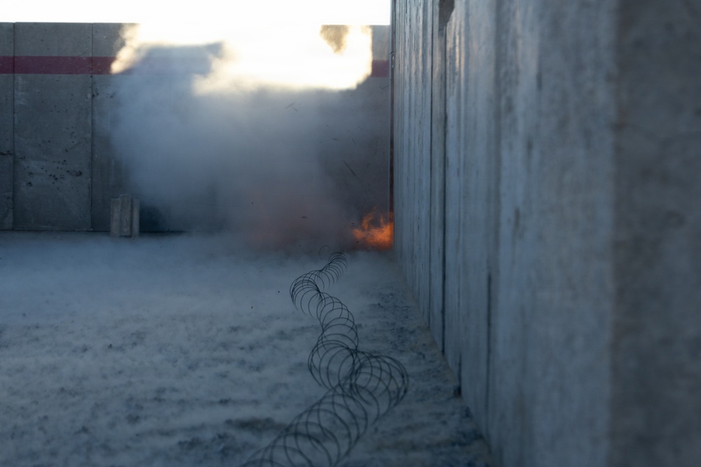 The image shows a narrow corridor between two concrete walls with razor wire coiled along the ground. In the background, there is visible fire and smoke billowing upward, suggesting an explosion or controlled burn. The scene has a tense, conflict-zone or security-perimeter aesthetic.