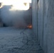 The image shows a narrow corridor between two concrete walls with razor wire coiled along the ground. In the background, there is visible fire and smoke billowing upward, suggesting an explosion or controlled burn. The scene has a tense, conflict-zone or security-perimeter aesthetic.